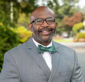 black man wearing glasses, grey suit coat, and a green bow tie. he is smiling.